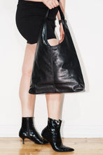 Load image into Gallery viewer, A person wearing black pointed-toe ankle boots and holding a large black leather hobo shoulder bag against a white wall and light wood floor.
